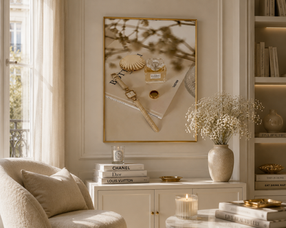 A framed wall art print with a gold border hangs above a white console table in a bright, elegant room. The print features a perfume bottle, shell, and jewelry on a beige background with soft botanical shadows.