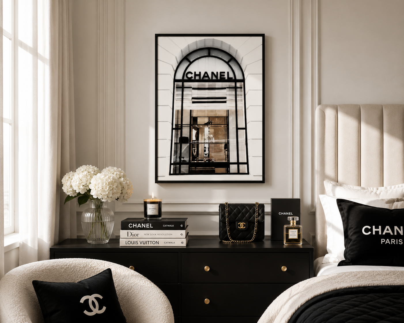 A framed black-and-white Chanel archway print hangs on a beige wall above a dark wood nightstand in a styled bedroom. The scene includes a tufted bed headboard, black bedding, Chanel and Louis Vuitton books, a quilted handbag, perfume, flowers, and pillows with Chanel branding.
