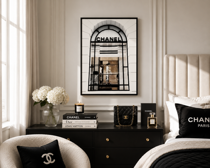 A framed black-and-white Chanel archway print hangs on a beige wall above a dark wood nightstand in a styled bedroom. The scene includes a tufted bed headboard, black bedding, Chanel and Louis Vuitton books, a quilted handbag, perfume, flowers, and pillows with Chanel branding.