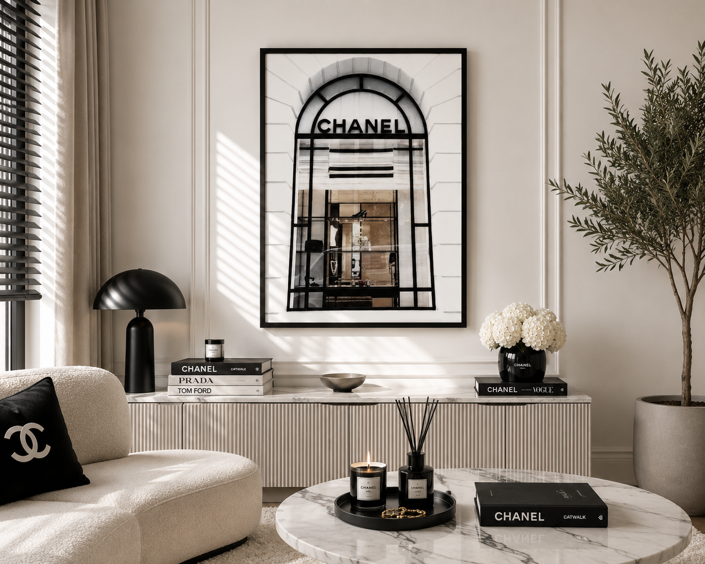 A framed black-and-white art print featuring a Chanel storefront archway hangs on a cream wall in a styled living room. The room includes a cream sofa with a black Chanel pillow, a white sideboard with books and decor, a marble coffee table, and a potted tree.