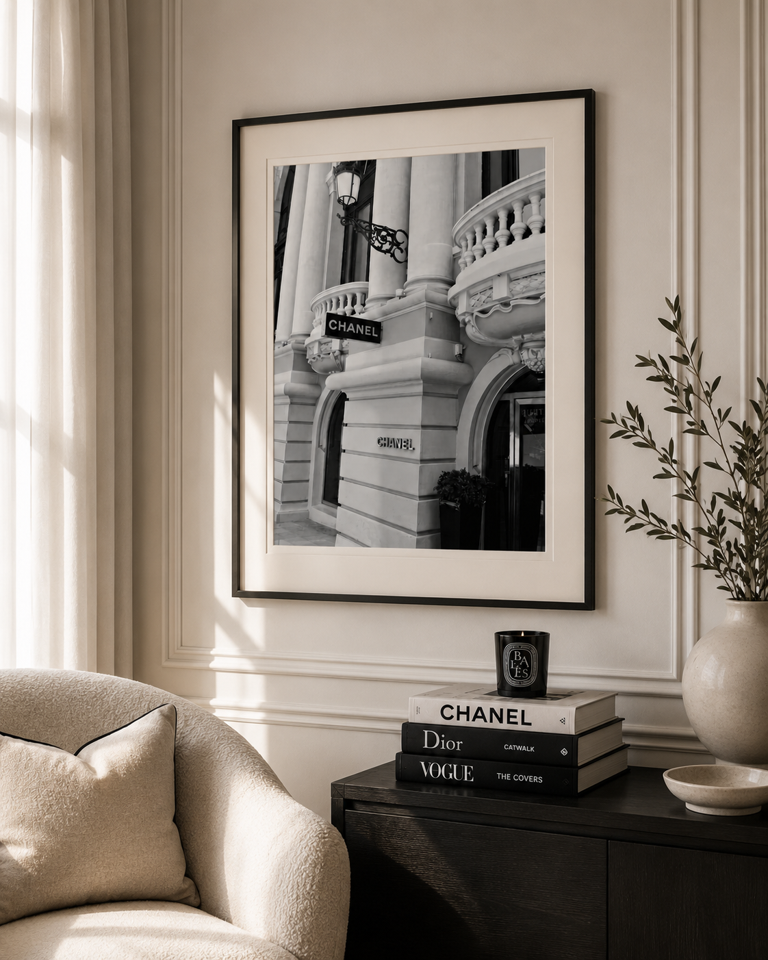 A framed black and white architectural photograph of a Parisian building with a visible Chanel sign, displayed on a beige wall above a dark sideboard with books and a vase in a styled interior.