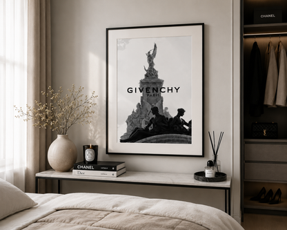A framed black and white Givenchy Paris poster hangs on a beige wall above a bedside table in a styled bedroom. The scene includes a bed, a vase with dried stems, books, a candle, a reed diffuser, and an open wardrobe with clothing and shoes.