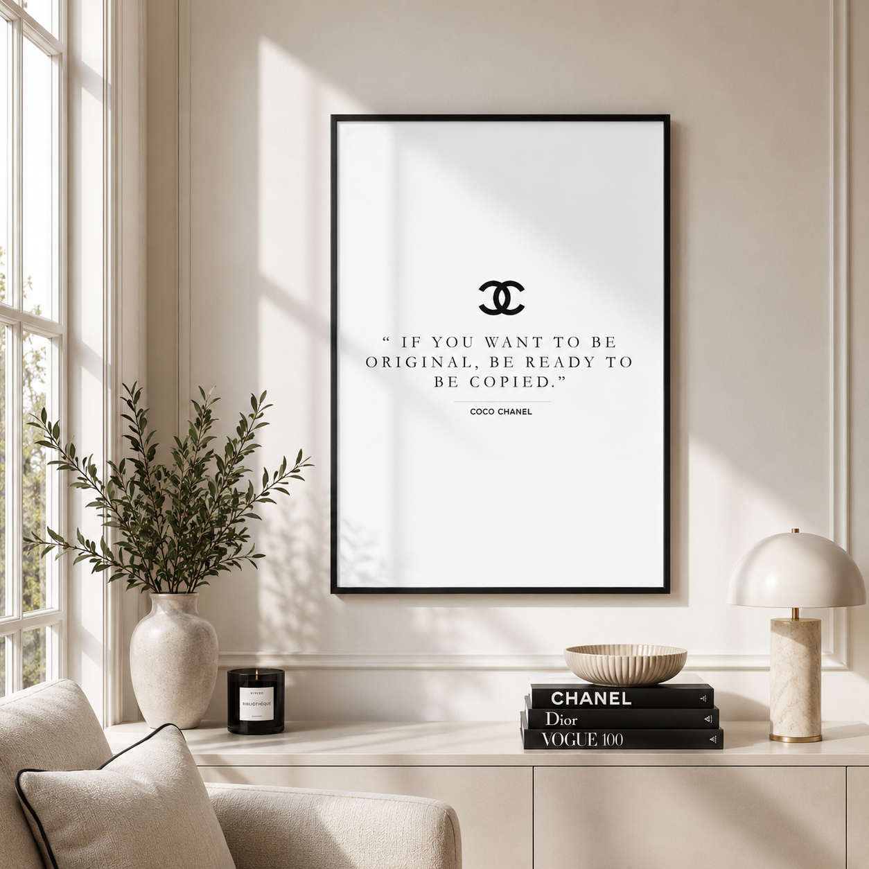 A framed wall art print featuring a black Chanel logo and the quote, 'IF YOU WANT TO BE ORIGINAL, BE READY TO BE COPIED.' by Coco Chanel, displayed in a bright, modern room above a console table with books, a lamp, and a vase.