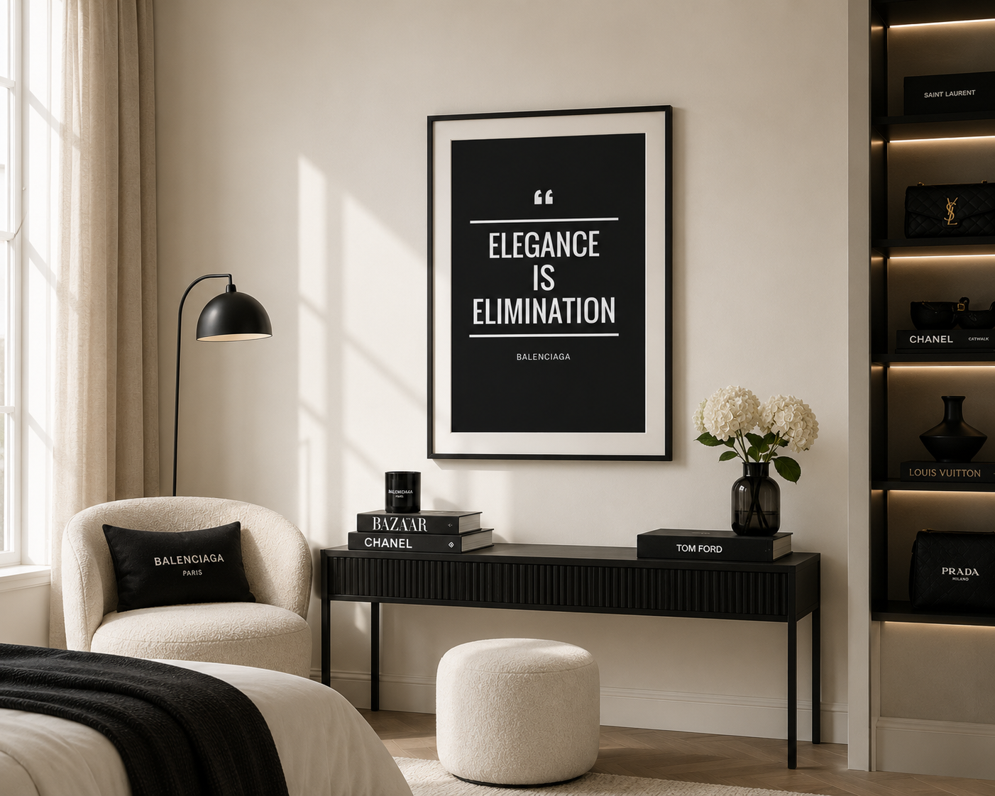 A framed black-and-white quote print reading “ELEGANCY IS ELIMINATION” hangs on a cream wall above a console table in a styled living room. The room includes a cream armchair with a Balenciaga pillow, a black floor lamp, a white ottoman, and shelving with luxury boxes and books.