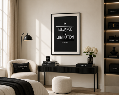 A framed black-and-white quote print reading “ELEGANCY IS ELIMINATION” hangs on a cream wall above a console table in a styled living room. The room includes a cream armchair with a Balenciaga pillow, a black floor lamp, a white ottoman, and shelving with luxury boxes and books.