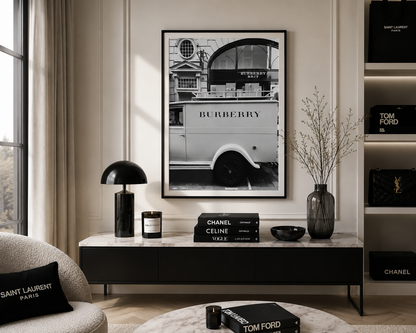 A framed black and white art print of a vintage Burberry double-decker bus hanging on a beige wall in a styled living room. The room features a black console table with a black dome lamp, a glass vase with dried branches, and books displaying Chanel, Celine, and Burberry branding.