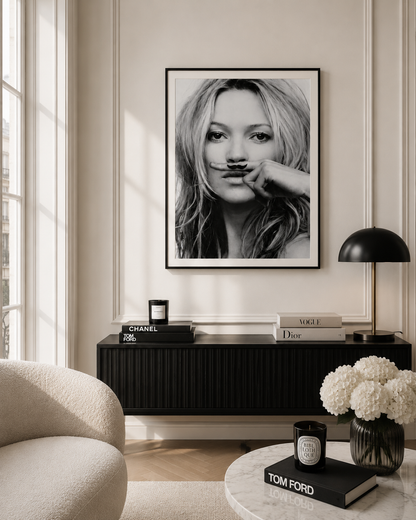 A black and white framed art print of a woman with a mustache hanging on a beige wall in a modern living room setting above a black sideboard.