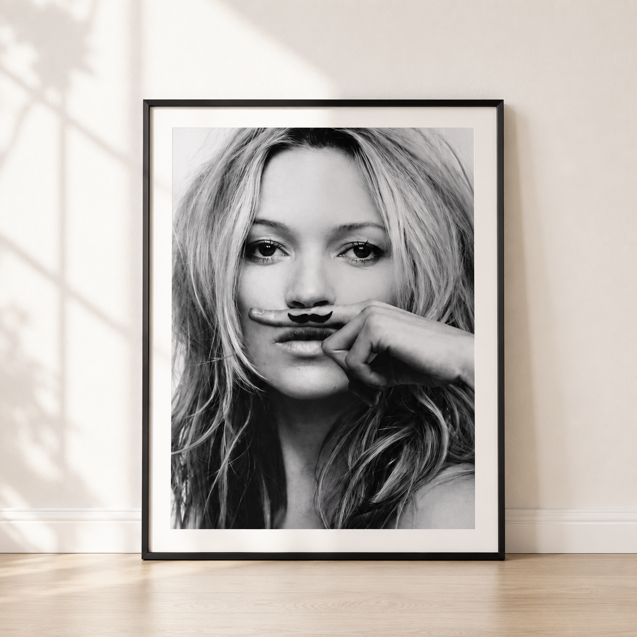 A framed black and white portrait print leaning against a light beige wall on a light wood floor. The print shows a woman with long hair holding a small object near her lips, and the artwork is enclosed in a black frame with a white mat.