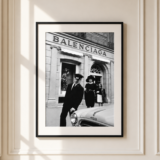 A framed black and white Balenciaga street photograph hanging on a cream-colored paneled wall with soft window shadows.