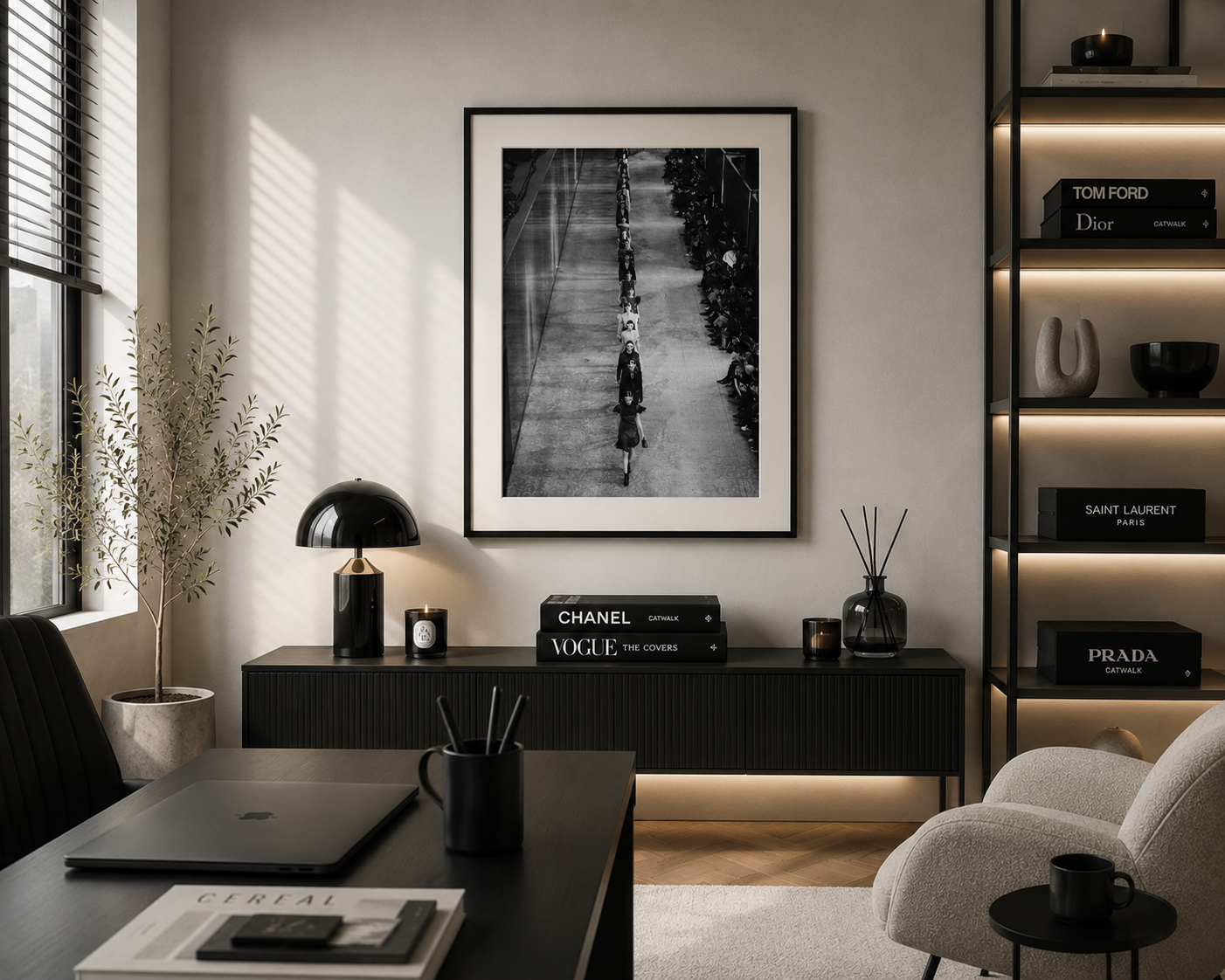A framed black and white art print hangs on a light beige wall in a modern, stylishly decorated office or living space. The room features a dark wood console table with a black desk lamp, a laptop on a dark table in the foreground, a light gray armchair, and a built-in shelving unit on the right with decorative books and vases.