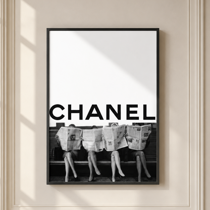A framed black and white poster featuring the word CHANEL in large bold letters above a black-and-white photograph of three women sitting on a bench with newspapers on their heads, displayed on a cream-colored wall with soft window shadows.