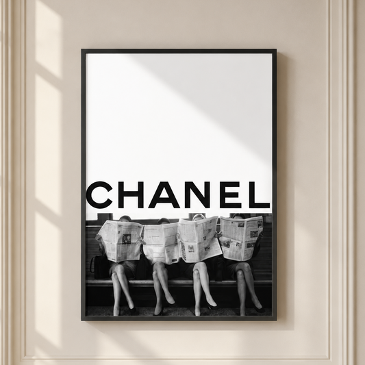 A framed black and white poster featuring the word CHANEL in large bold letters above a black-and-white photograph of three women sitting on a bench with newspapers on their heads, displayed on a cream-colored wall with soft window shadows.