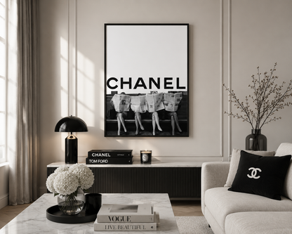 A framed black and white Chanel poster hangs on a light beige wall in a styled living room. The room features a white sofa with a black Chanel throw pillow, a marble coffee table with books, and a black table lamp.