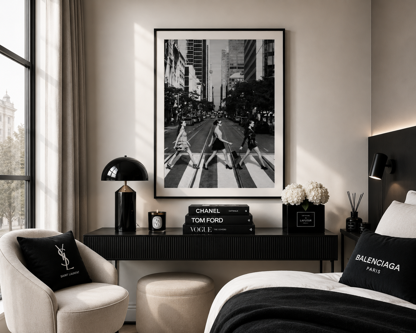 A framed black and white street photograph hanging on a beige wall above a dark console table in a modern bedroom. The room includes a cream armchair with a black pillow, a bed with a black Balenciaga pillow, books, and lamps.