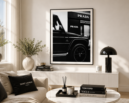 A framed black and white art print of a Mercedes-Benz G-Class vehicle with the Prada logo is displayed on a wall in a modern, sunlit living room setting.