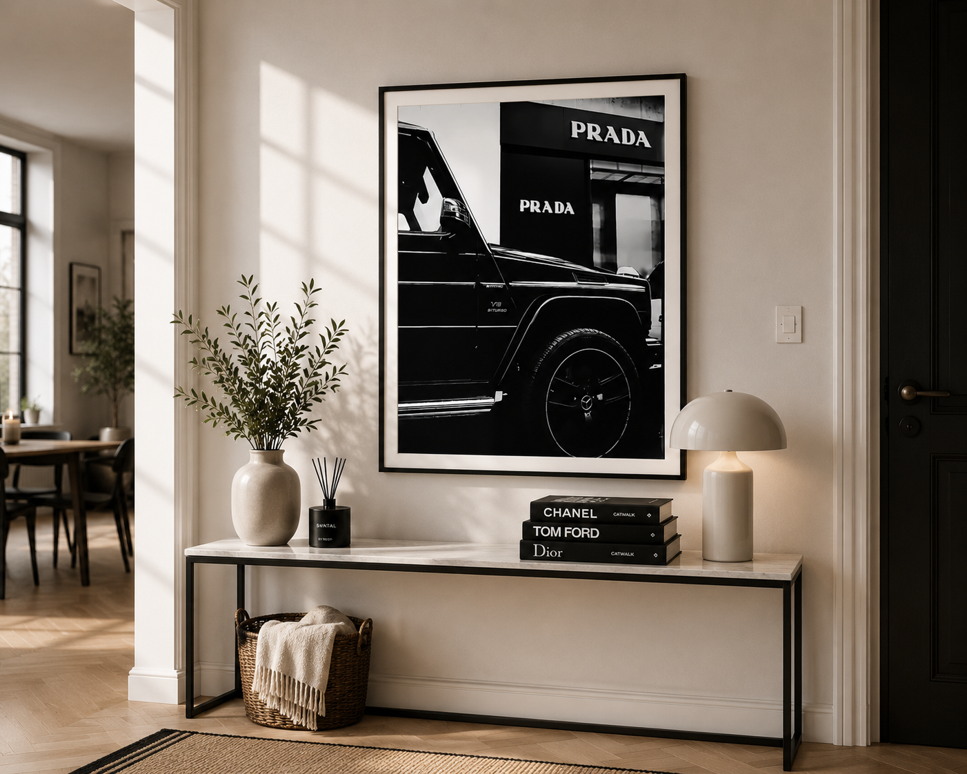 A framed black-and-white wall art print featuring a luxury car and the PRADA logo, displayed in a modern, sunlit living room above a console table with decorative books and a vase.
