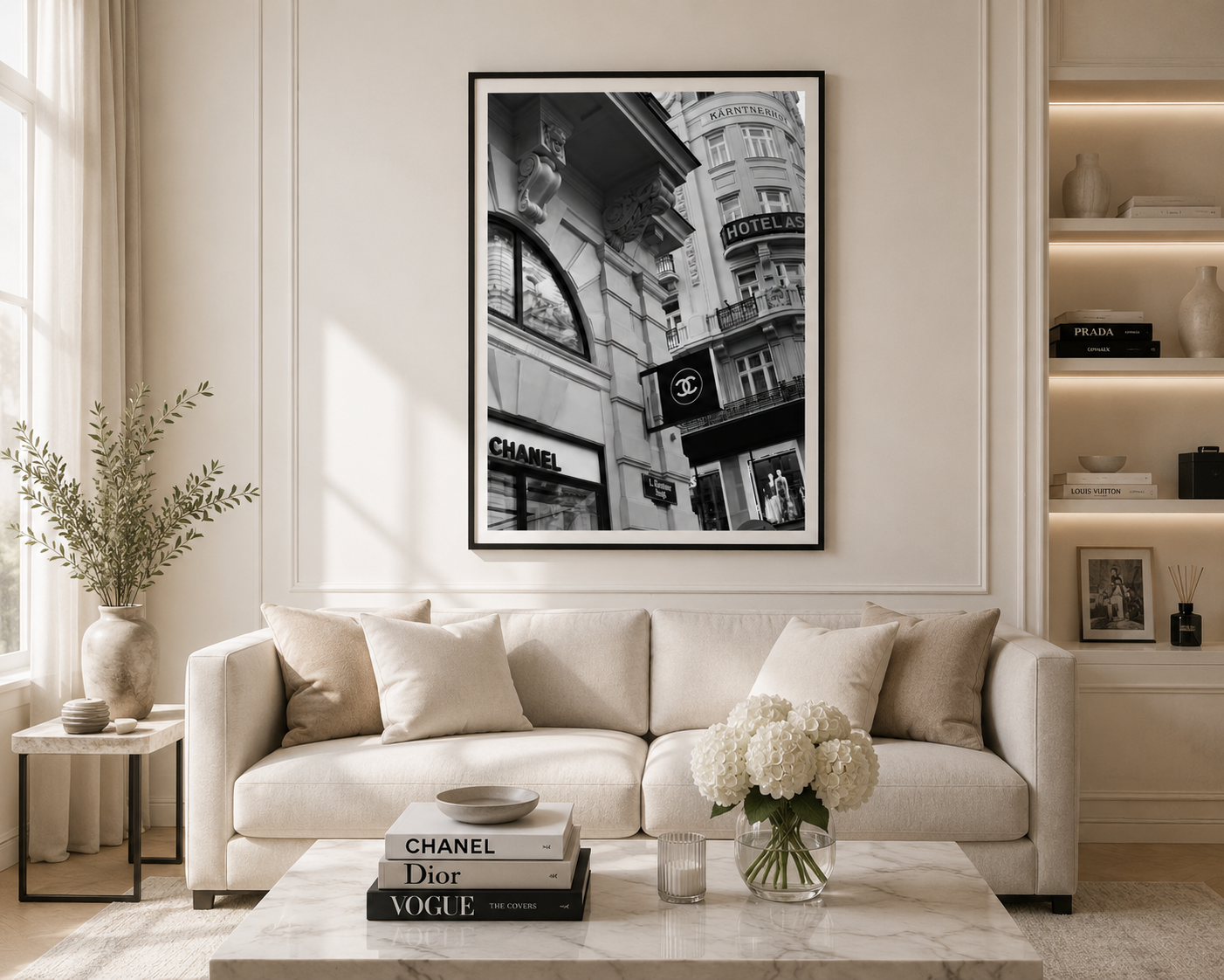 A framed black and white architectural street art print hangs on a cream wall above a beige sofa in a modern, sunlit living room. The print features classic European buildings with visible Chanel and Dior storefronts.