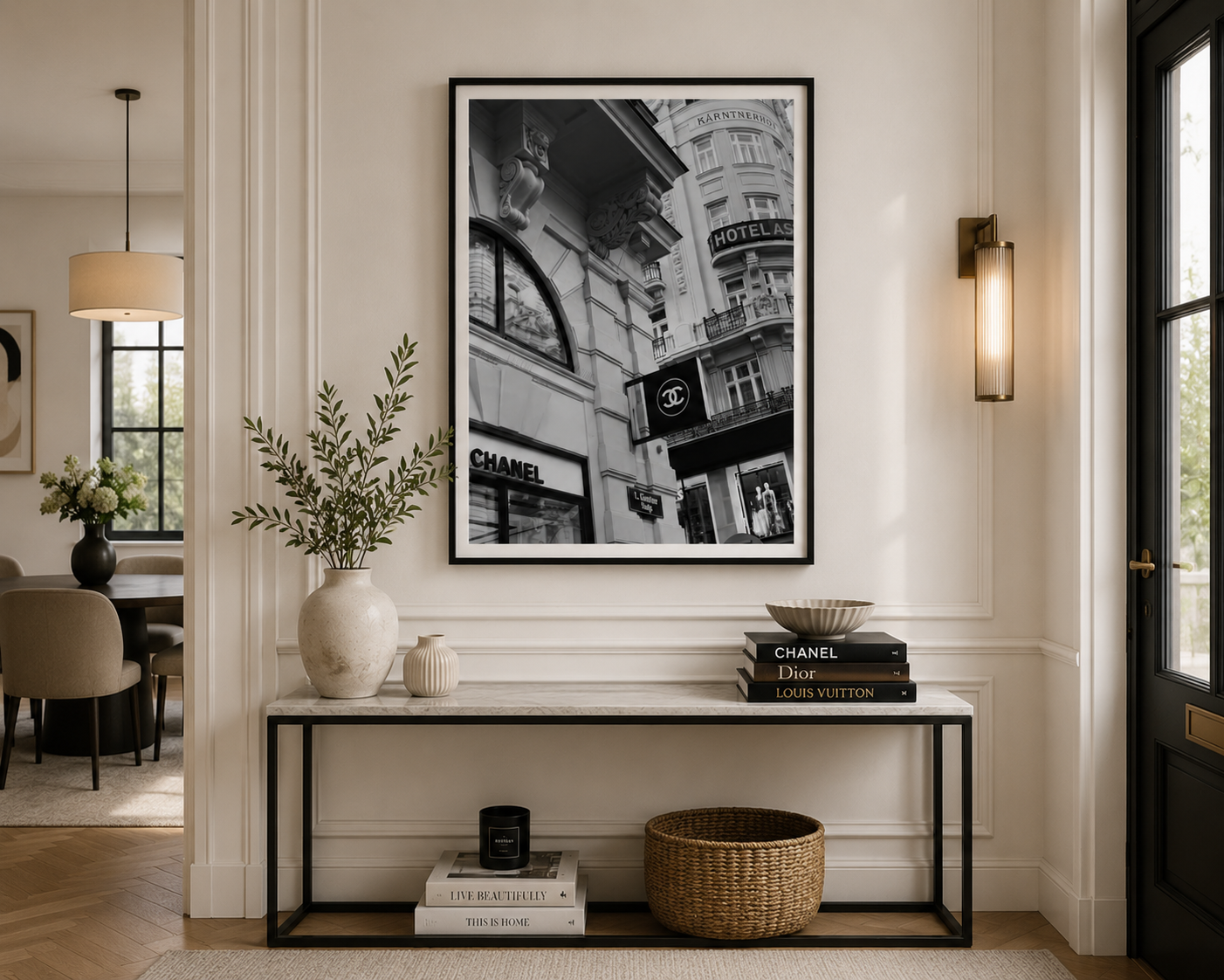 A large black and white framed art print depicting a Parisian street scene with iconic Chanel and Louis Vuitton storefronts, displayed on a white wall above a console table in a beautifully styled modern entryway.