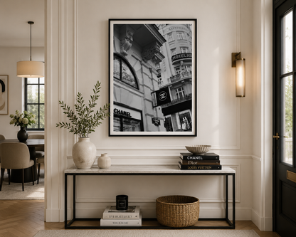 A large black and white framed art print depicting a Parisian street scene with iconic Chanel and Louis Vuitton storefronts, displayed on a white wall above a console table in a beautifully styled modern entryway.
