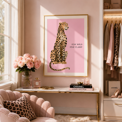 A framed art print featuring a pink background with a spotted cheetah illustration and the text 'STAY WILD STAY CLASSY' hangs on a pink wall in a styled room. The scene includes a blush pink velvet chair with a leopard-print pillow, a console table with flowers and books, and a clothing rack with Chanel and Louis Vuitton items.