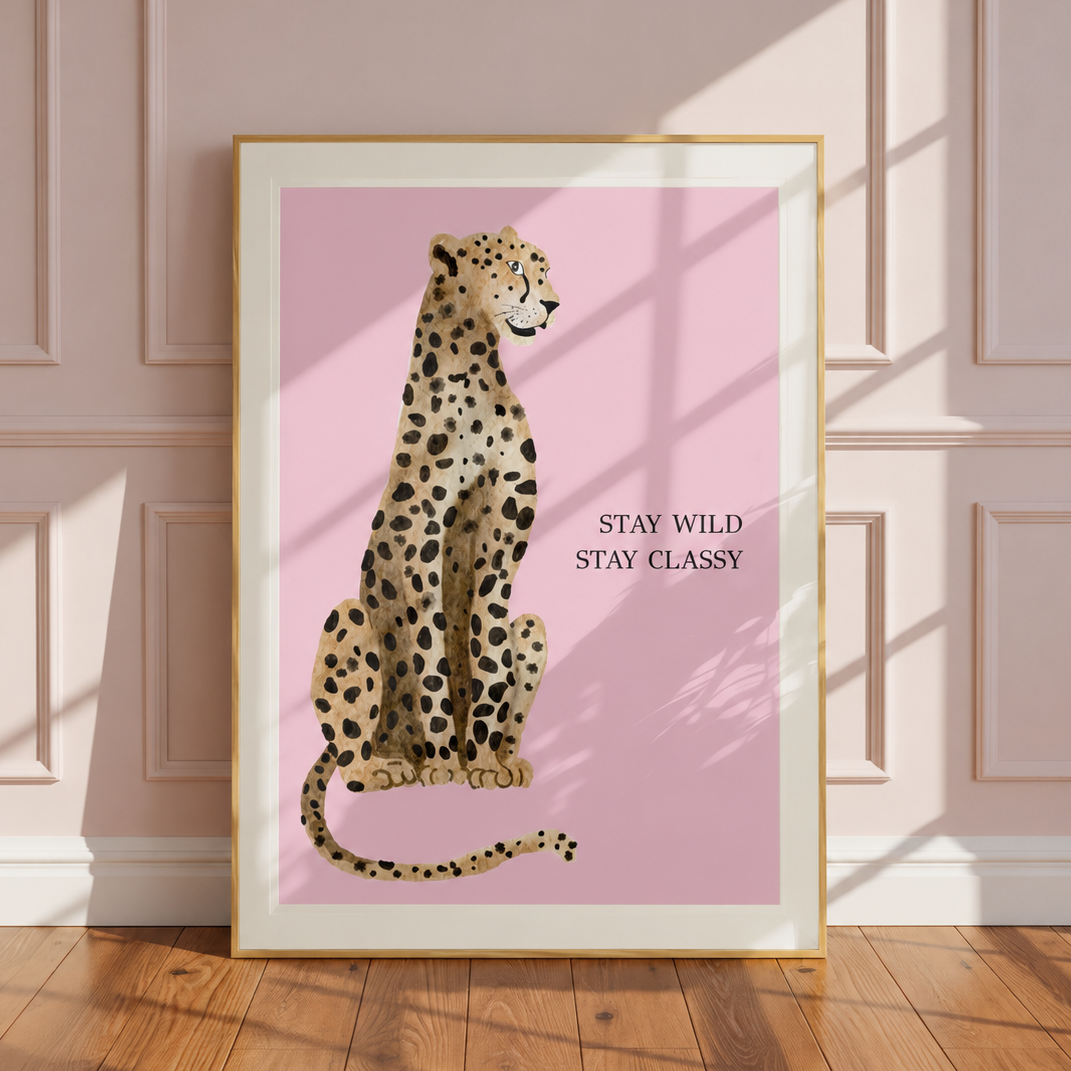 A framed art print featuring a seated cheetah on a pink background with the text 'STAY WILD STAY CLASSY', leaning against a light pink paneled wall on a wooden floor.