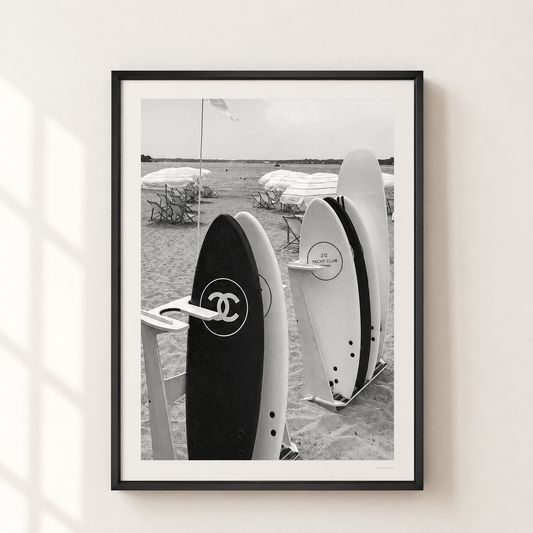 A framed black and white photographic print of surfboards on a beach, featuring a Chanel logo on one board, displayed on a light beige wall with soft window shadow overlays.