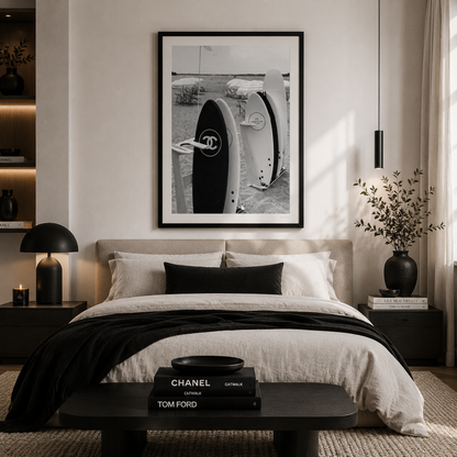 A styled bedroom interior with a large black-and-white framed wall print above a bed. The print shows a close-up of a circular object with a double-C logo, suggesting Chanel branding.