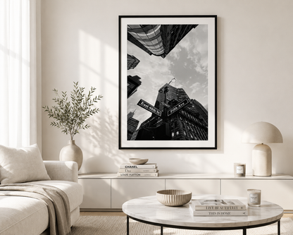 A framed black and white urban photograph of skyscrapers and street signs hangs on a light beige wall in a modern living room setting with a sofa, coffee table, and lamp.