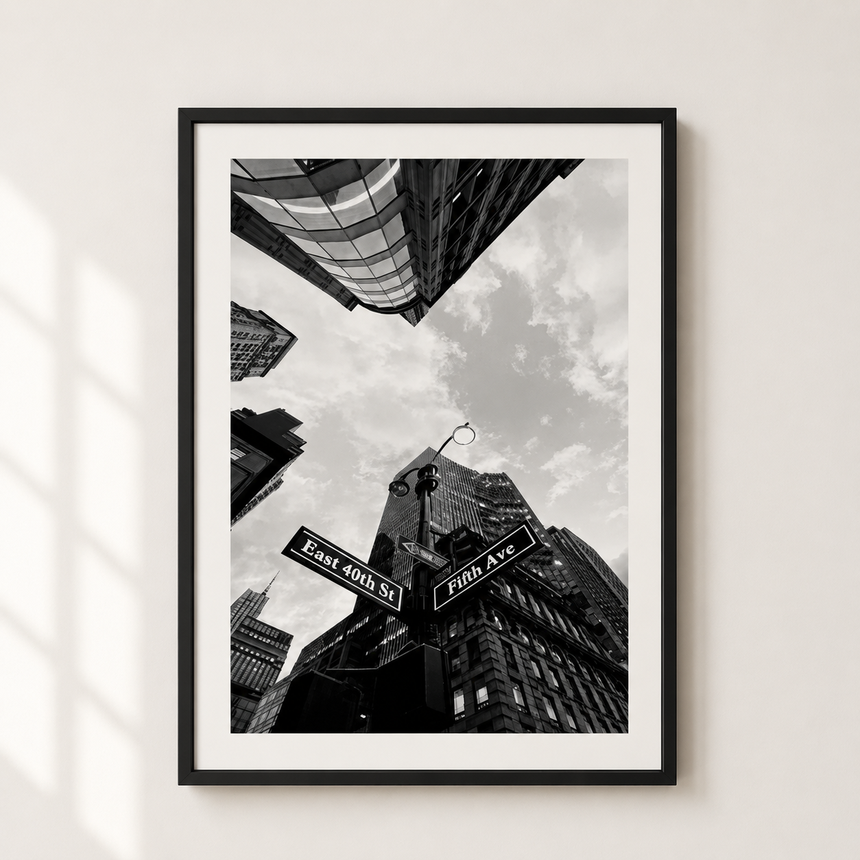 A framed black and white street art print depicting upward-looking skyscrapers and street signs reading East 40th St and Fifth Ave, hung on a light beige wall with soft window shadows.