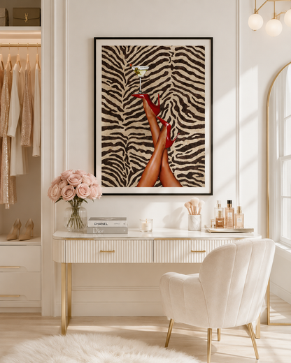 A framed art print featuring a zebra-stripe pattern background and a pair of legs in red high heels holding a cocktail, displayed on a white wall above a vanity desk in a styled room.