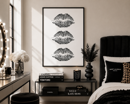 A framed wall art print featuring three black lip kiss marks on a white background, styled in a modern bedroom setting next to a bed and vanity table.