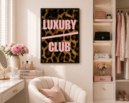 A framed wall art print featuring a leopard print background with the words 'LUXURY CLUB' in pink, displayed in a styled room with a cream chair, vanity, and shelving.