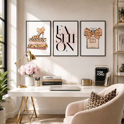 Three framed fashion-themed art prints hang on a beige wall above a white desk in a styled home office. The prints feature a leopard-print high-heel shoe, the word 'FASHION' in large black letters, and a Chanel perfume bottle with a leopard bow.