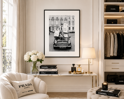 A framed black and white art print of a vintage car with a woman on top, displayed on a cream wall in a styled room with a sofa, console table, and shelving.