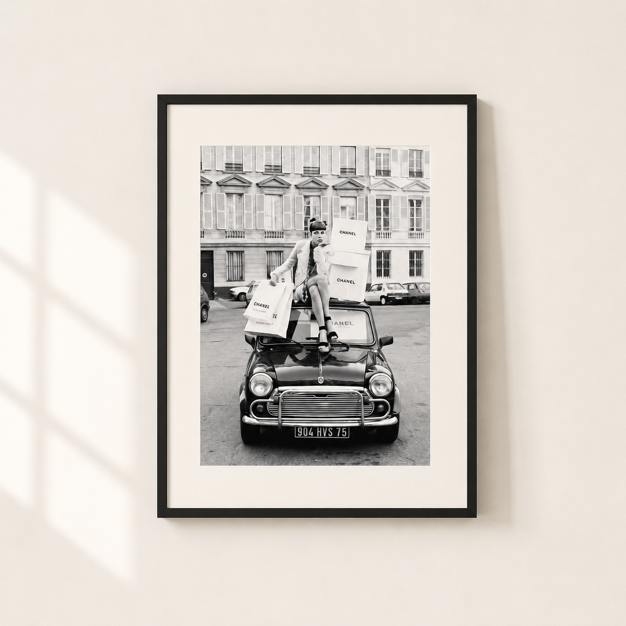A framed black and white art print depicting a vintage car with a person carrying shopping bags, mounted on a light beige wall with soft window shadows.