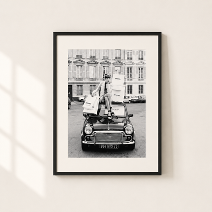 A framed black and white art print depicting a vintage car with a person carrying shopping bags, mounted on a light beige wall with soft window shadows.