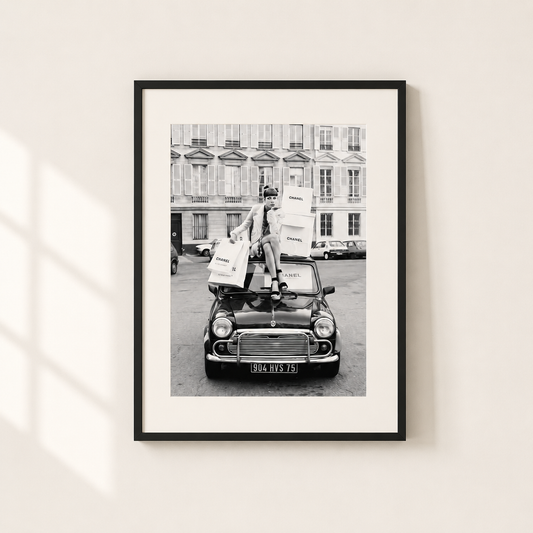 A framed black and white art print depicting a vintage car with a person carrying shopping bags, mounted on a light beige wall with soft window shadows.