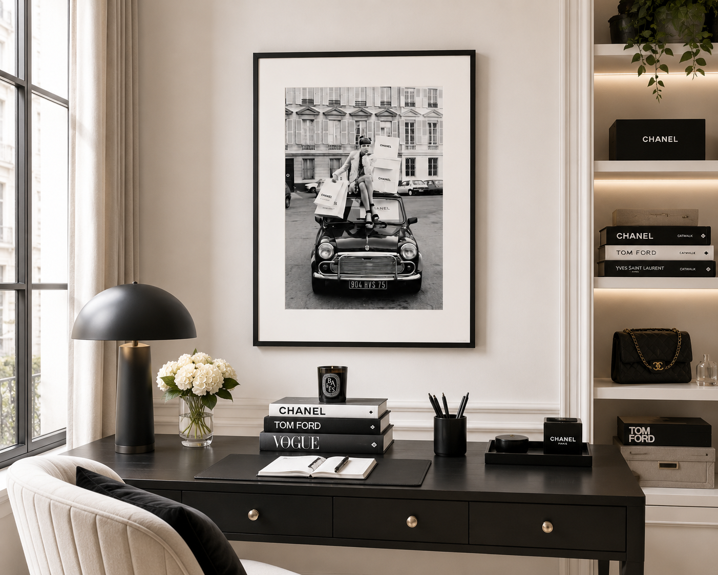 A black and white framed art print hangs on a light beige wall above a dark wood desk in a styled home office. The desk is decorated with a black dome lamp, a vase of white flowers, a notebook, and books, including Chanel and Tom Ford editions.