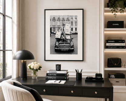 A black and white framed art print hangs on a light beige wall above a dark wood desk in a styled home office. The desk is decorated with a black dome lamp, a vase of white flowers, a notebook, and books, including Chanel and Tom Ford editions.