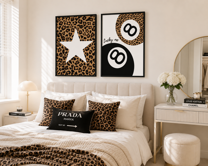A styled bedroom interior with two framed wall art prints above a bed. The prints feature leopard print backgrounds: one with a large white star and the other with a black-and-white billiard ball motif and the words 'Lucky me.
