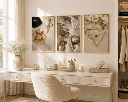 Three framed wall art prints with gold borders hang above a white vanity desk in a bright, elegant room. The prints feature luxury-themed imagery including Chanel bottles, seashells, and gold accessories.