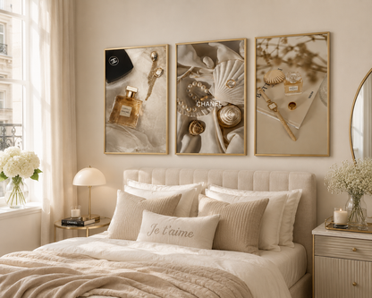 Three framed vertical wall art prints with gold frames hang above a bed in a softly lit beige bedroom. The prints feature luxury-inspired imagery including perfume bottles, shells, jewelry, and gold-toned objects on neutral backgrounds.