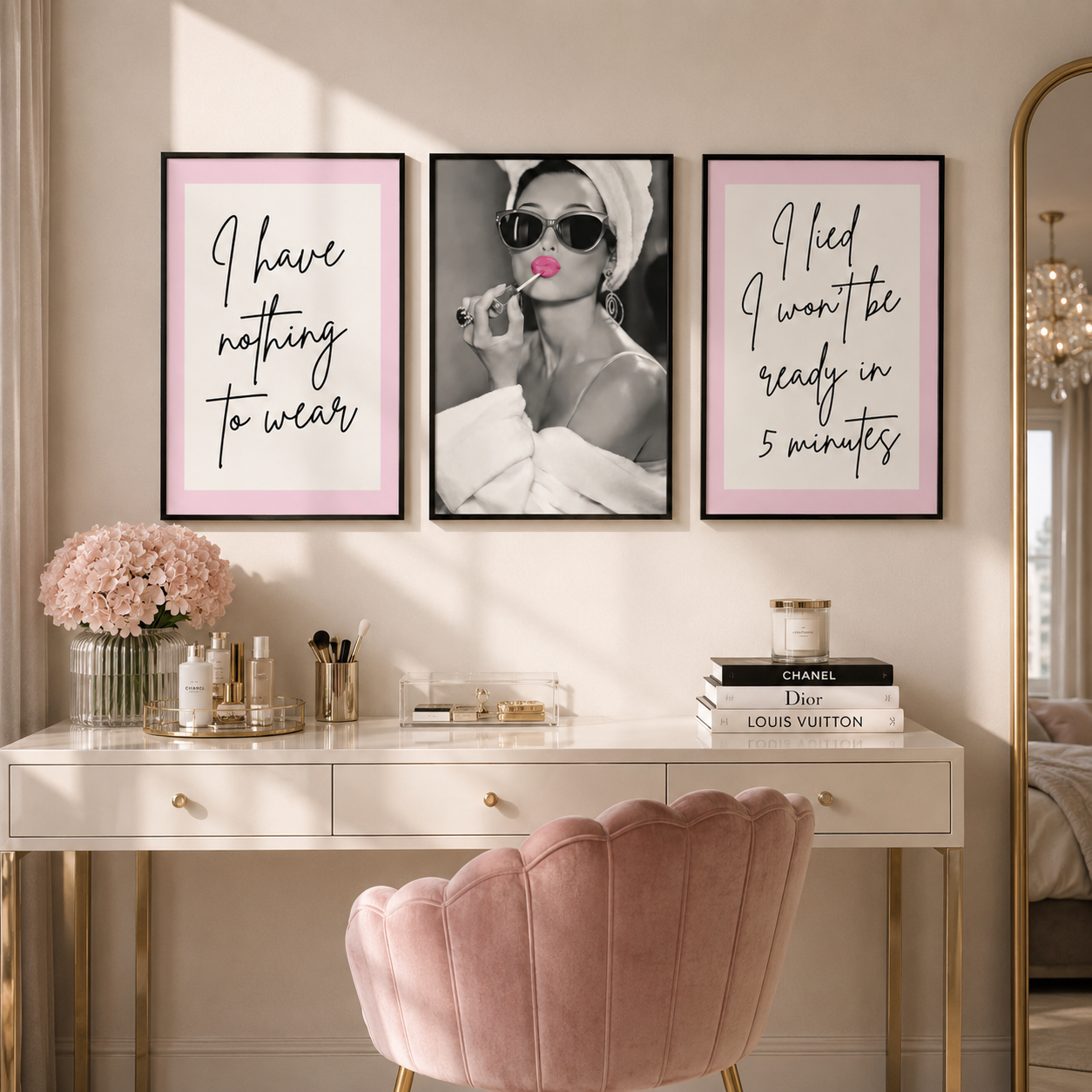 Three framed wall art prints hang above a white vanity desk in a styled bedroom. The left print says 'I have nothing to wear' on a pale pink background, the center print is a black-and-white fashion photo of Audrey Hepburn with pink lipstick and sunglasses, and the right print says 'I lied I won't be ready in 5 minutes' on a pale pink background.