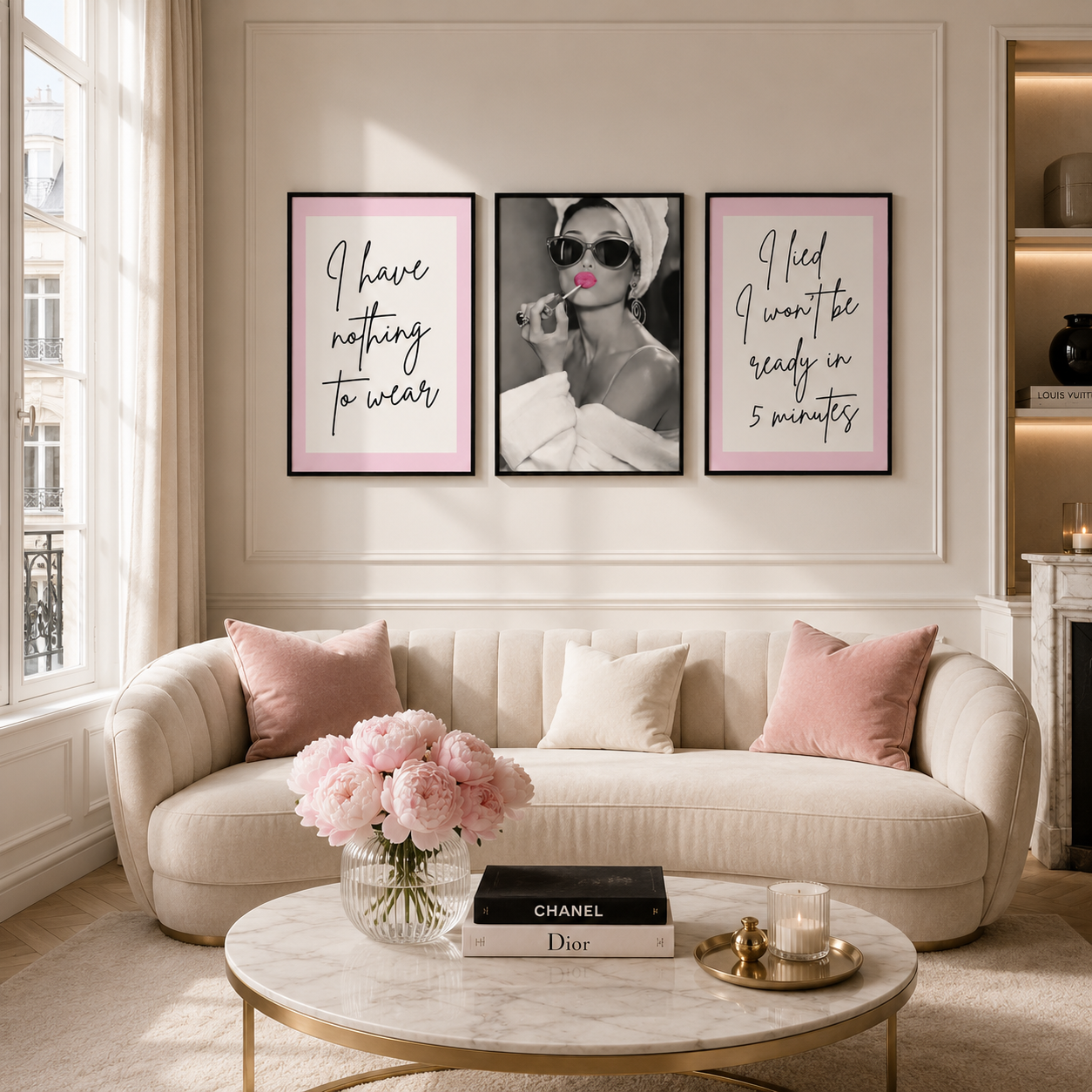 Three framed wall art prints with pink borders hang above a cream sofa in a bright living room. The center print shows a black-and-white portrait of Audrey Hepburn in a headscarf applying pink lipstick while the two side prints contain cursive text in black.