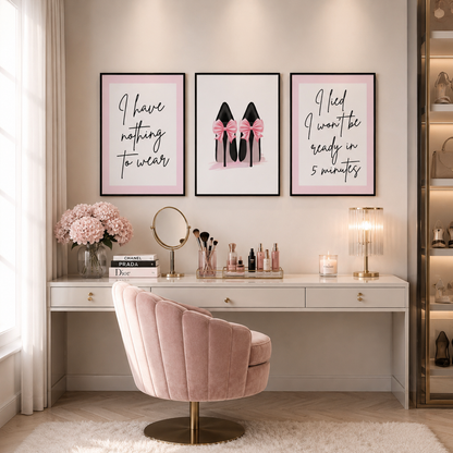 A styled vanity area with three framed motivational and fashion prints above a white desk and blush pink velvet swivel chair. The center print shows black high heels with pink bows, while the side prints contain handwritten text.