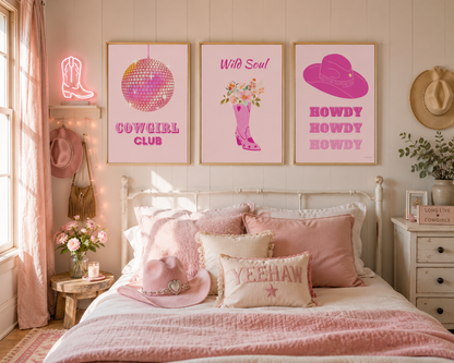 A styled bedroom scene with three framed western-themed art prints above a bed. The prints read 'Cowgirl Club' with a disco ball graphic, 'Wild Soul' with a pink boot and flowers, and 'Howdy Howdy' with a pink hat.