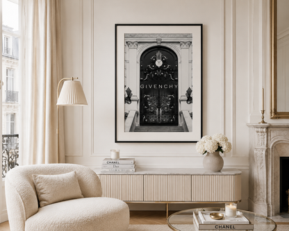 A framed black and white art print of a Givenchy door hanging on a cream paneled wall in a styled living room with a beige armchair, console table, and fireplace.