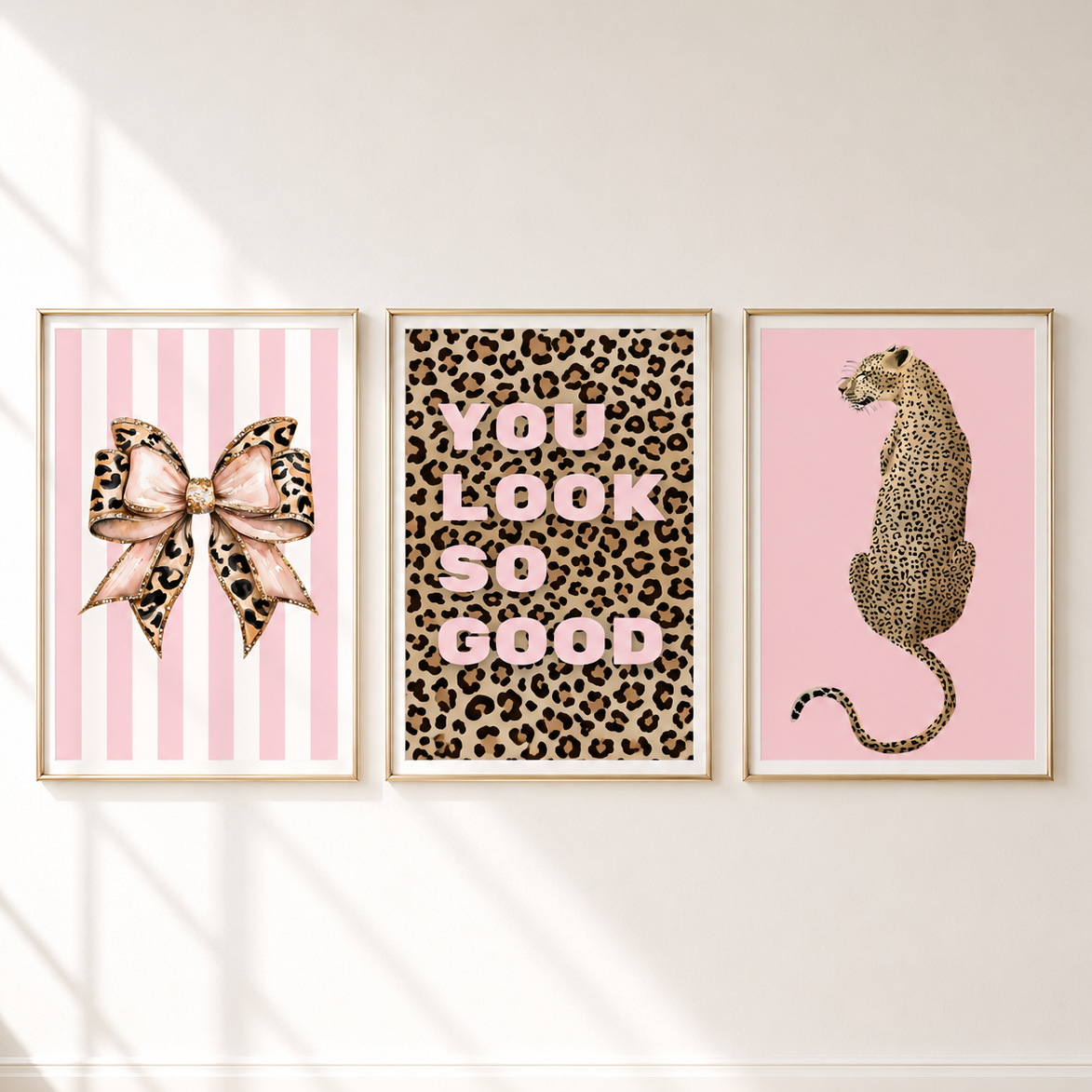 Three framed wall art prints displayed side by side on a white wall. The left print shows a pink striped background with a leopard-print bow; the middle print has a leopard-print background with pink text reading 'YOU LOOK SO GOOD'; the right print shows a pink background with a leopard sitting in profile.