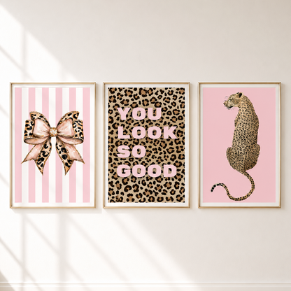 Three framed wall art prints displayed side by side on a white wall. The left print shows a pink striped background with a leopard-print bow; the middle print has a leopard-print background with pink text reading 'YOU LOOK SO GOOD'; the right print shows a pink background with a leopard sitting in profile.