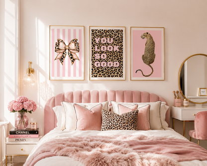 A styled bedroom scene with three framed wall art prints above a pink tufted bed. The prints feature pink-and-white stripes with a leopard-print bow, pink text reading 'YOU LOOK SO GOOD' over leopard print, and a pink leopard-print cheetah.